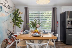 A dining area with a wooden table, white chairs, a map mural, and a window with blinds. - The Hideaway~Pool~Pickleball~Putt-Putt~Kayaks! (Marathon)