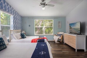 Ein Schlafzimmer mit zwei Betten, einem Deckenventilator, einem Fernseher, einem Schminktisch und einem Fenster mit Jalousien.