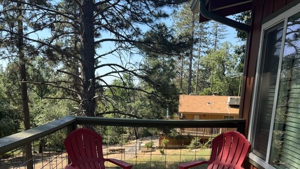 Una terraza de madera con dos sillas Adirondack rojas, una mesa con dos platos de comida, y una vista de árboles y un edificio al fondo.