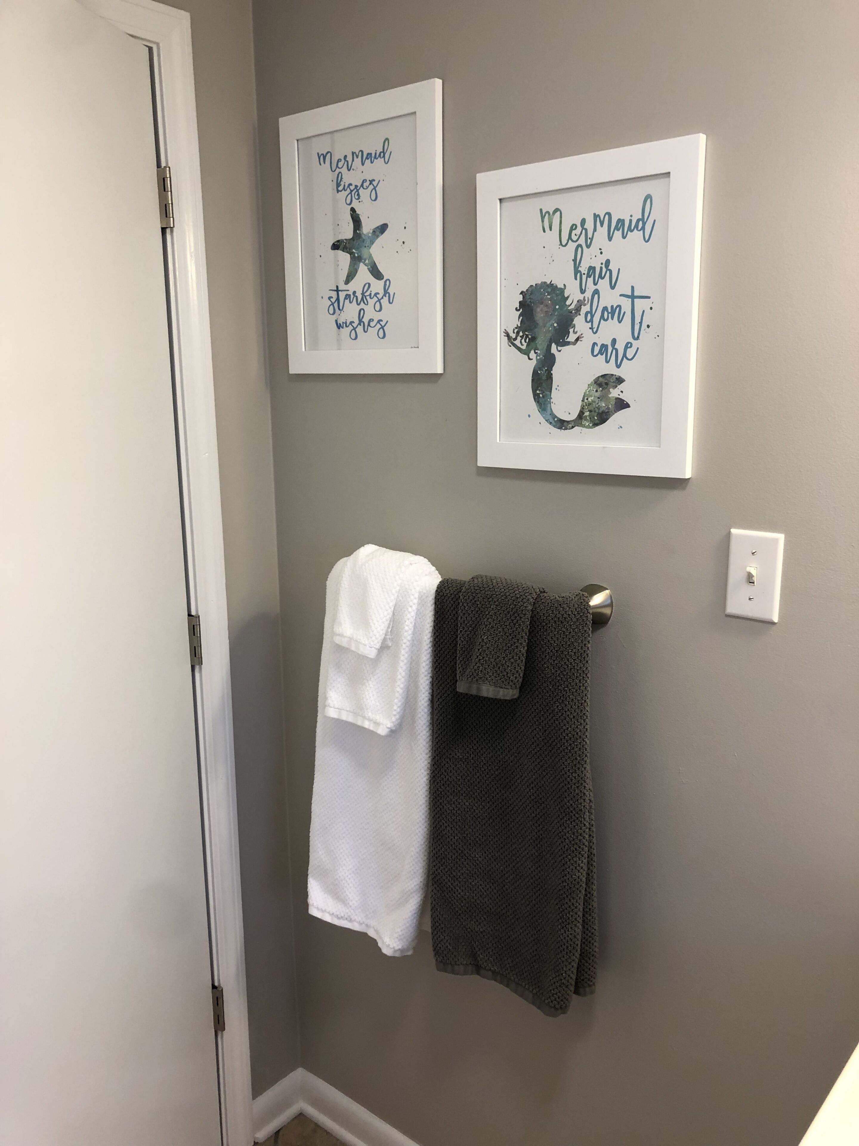 A bathroom with two framed wall art pieces depicting mermaids and sea creatures, a white towel, and a gray towel hanging on a towel rack.