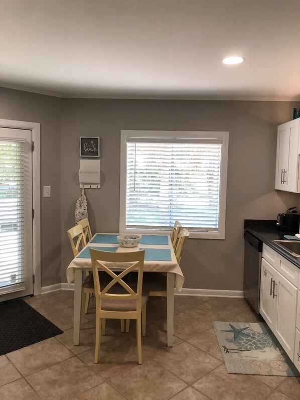 Un comedor con una mesa dispuesta para cuatro, una ventana con persianas y una zona de cocina con armarios y un lavabo.
