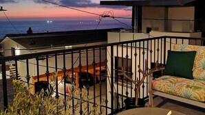 A balcony with a fire pit, outdoor seating, and a view of the ocean at sunset.
