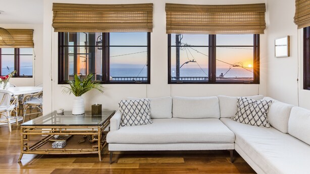 A modern living room with a white sofa, two windows with bamboo blinds, a glass coffee table, and a view of the sunset.