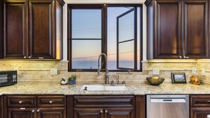 A kitchen with dark wood cabinets, a granite countertop, a stainless steel dishwasher, and a window with an open view.