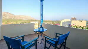 Un balcon avec deux chaises bleues et une petite table, d’où l’on peut admirer un paysage pittoresque composé de collines et de bâtiments.