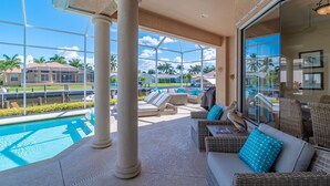 Un espace aménagé au bord de la piscine, avec des chaises longues, une table à manger et une grande fenêtre offrant une vue sur les palmiers et les maisons.