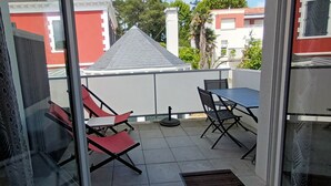 A balcony with red and black chairs, a table, and a view of neighboring buildings. - Apartment T2 ANGLET (Anglet)