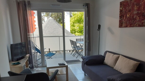 A living room with a sofa, a TV, a coffee table, and a chair. There is a balcony with a view of trees and a building. - Apartment T2 ANGLET (Anglet)