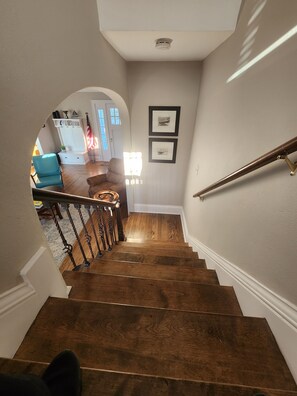 Uma escada de madeira com corrimão, que leva a uma sala de estar com sofá, mesa de centro e quadros nas paredes.