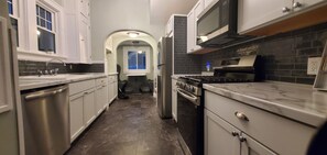 Cozinha com armários brancos, painel de azulejos pretos, eletrodomésticos de aço inoxidável e bancada de mármore.