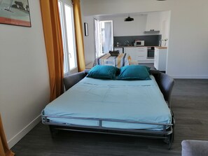 A single bed with teal bedding in a room with a dark wooden floor, a window with curtains, and a kitchenette in the background.
