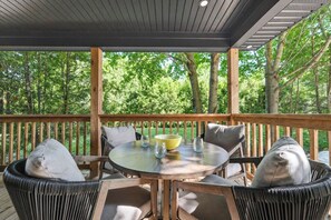 A covered outdoor seating area with a round table, chairs, and a view of a wooded area.