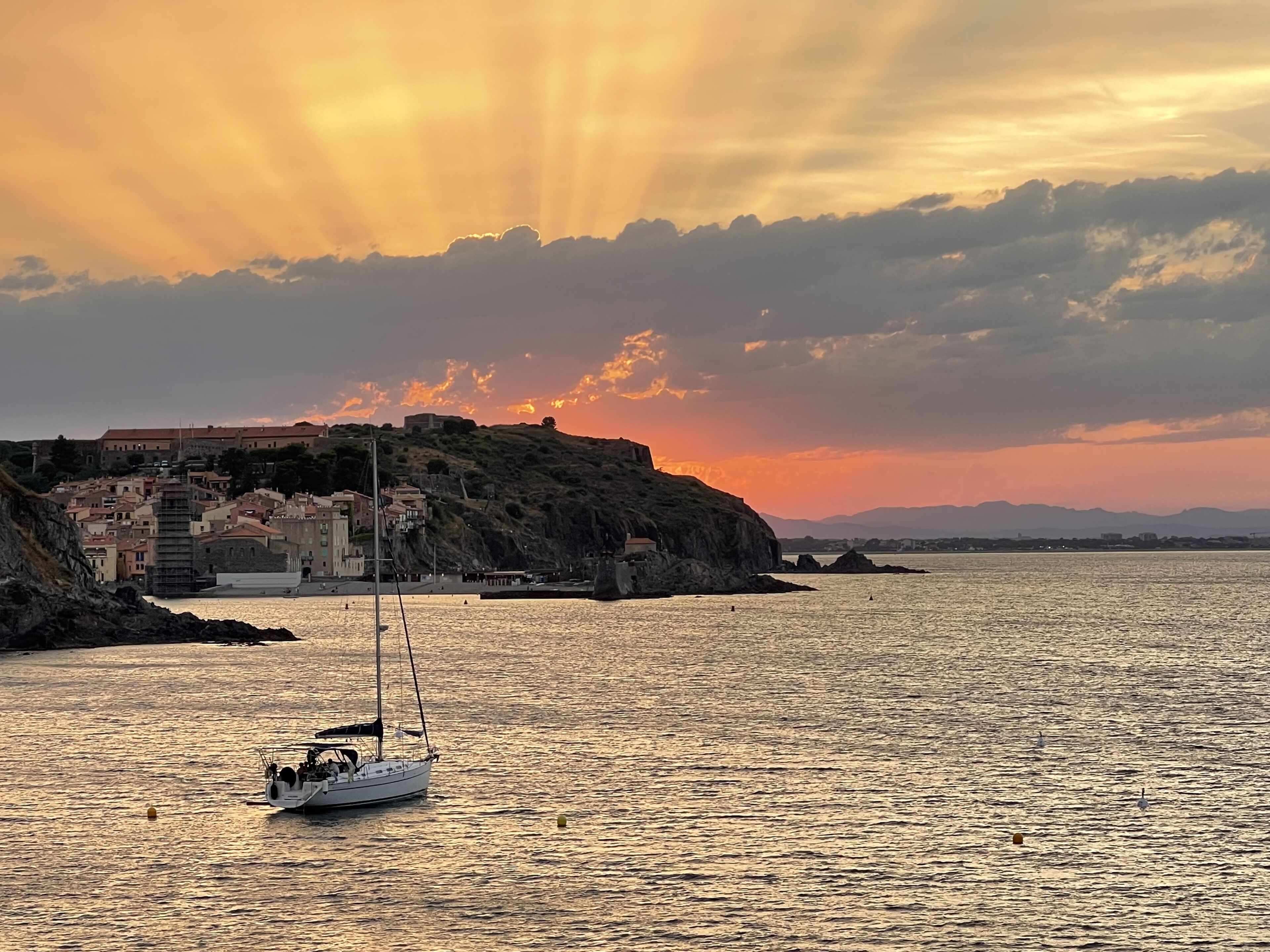 A coastal scene with a sailboat on calm waters, a historic building on a cliff, and a sunset sky.