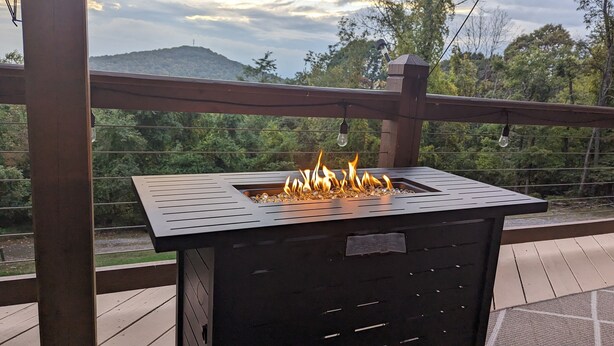 Un barbecue extérieur en métal, avec des flammes, installé sur une terrasse en bois, avec en arrière-plan une colline boisée.