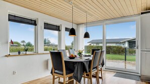 Uma área de refeiç ões com uma mesa, cadeiras e luminárias pendentes. Há janelas amplas com persianas, vista para o exterior e uma porta que dá para uma varanda.