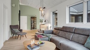 Uma sala de estar moderna com um sofá cinzento, uma mesa de centro redonda com livros e uma tigela de fruta, uma poltrona cinza e um aparador de vidro com objetos decorativos.