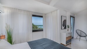 Un dormitorio con cama, mesita de noche, silla, ventana con vistas y una obra de arte en la pared.