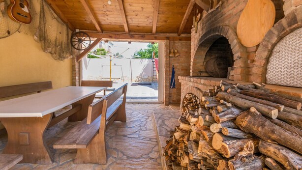 Un intérieur rustique avec un sol en pierre, des bancs en bois, une table, une cheminée avec des bûches et une vue sur l’extérieur.