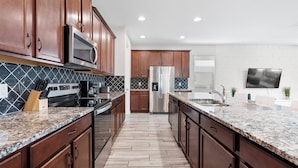 A modern kitchen with wooden cabinets, a stainless steel refrigerator, a microwave, and granite countertops. - 9BR house with private pool in Solara Resort (Kissimmee)