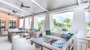 A covered outdoor lounge area with multiple seating options, ceiling fans, and large windows offering views of palm trees. - 9BR house with private pool in Solara Resort (Kissimmee)