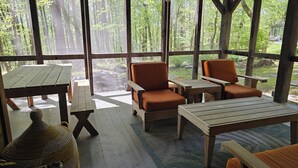 A wooden deck with a table and two orange chairs, a woven basket, and a view of a forest through large windows.
