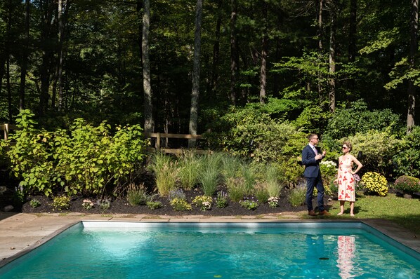 A pool with a wooden deck and lush greenery in the background.