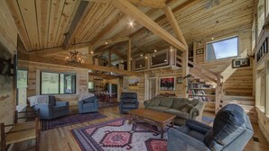 Ein großzügiges Blockhaus-Interieur mit hohen Decken, Holzbalken, einer zentralen Sitzgruppe mit Sofas und Sesseln, einem großen Teppich und einer Treppe, die in einen oberen Bereich führt.