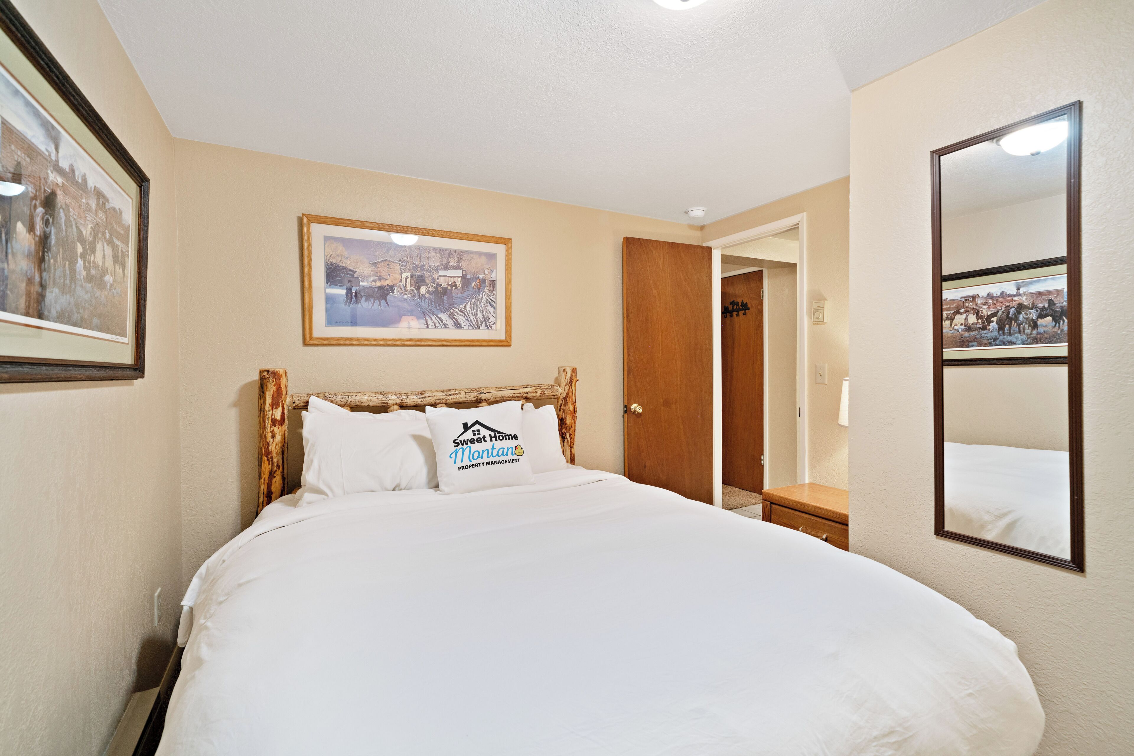 A hotel room with a large bed, a wooden headboard, a framed picture on the wall, a mirror, and a door leading to another room.