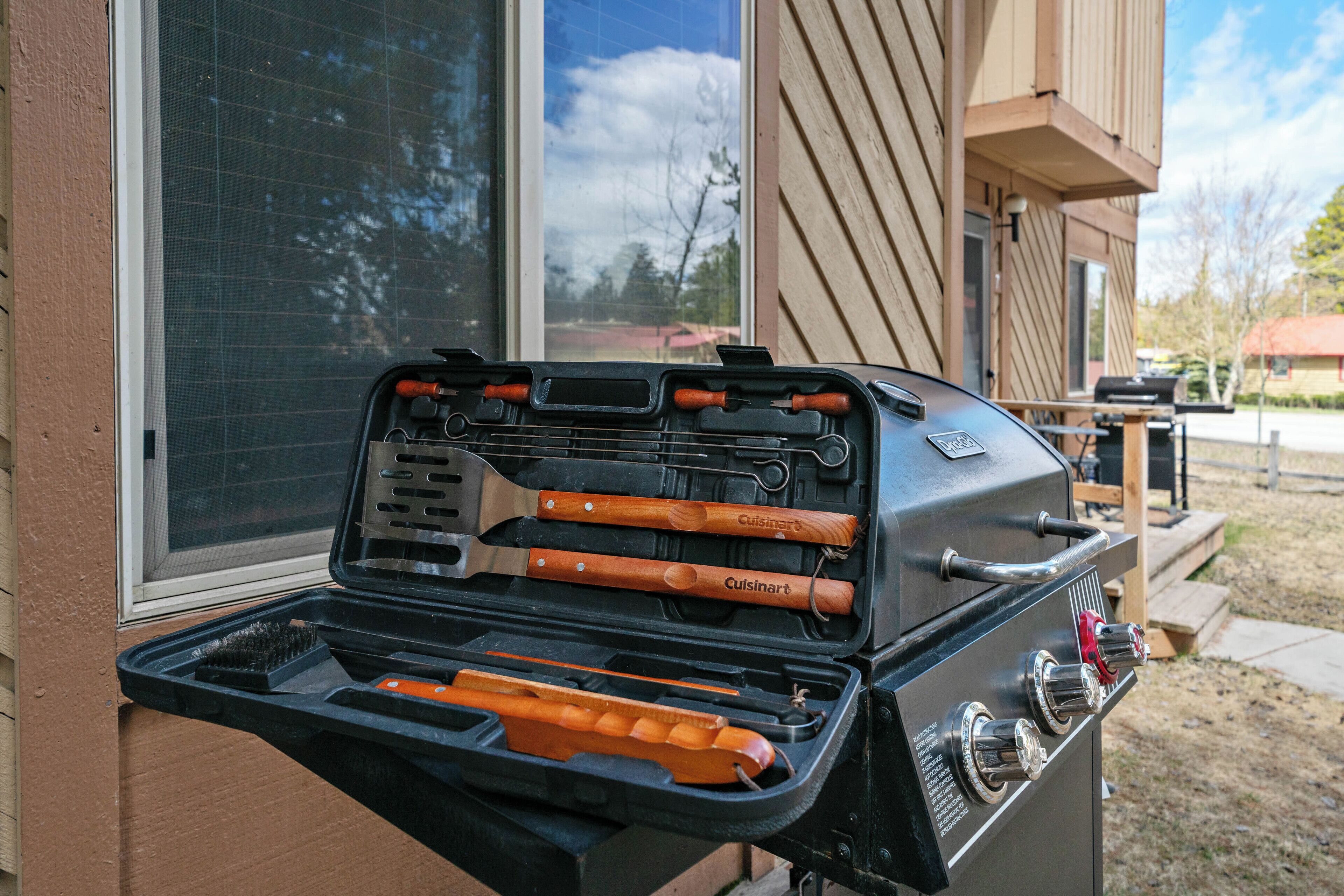 Un barbecue avec des ustensiles et un manche en bois, placé à l’extérieur d’un bâtiment doté d’une fenêtre et d’une terrasse.