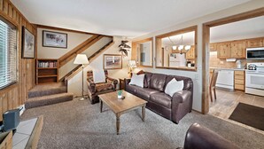 A living room with a brown leather sofa, a wooden coffee table, a bookshelf, a lamp, and a staircase leading to a kitchen area. - Trapper 8 (West Yellowstone)