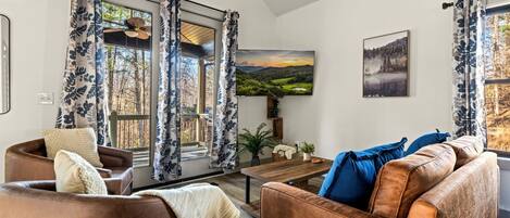 Una sala de estar con un sofá de cuero marrón, una mesa de centro de madera y un ventanal con cortinas florales azules. En la pared, hay un cuadro de un paisaje.