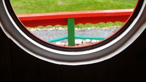 Blick durch ein rundes Fenster mit rotem Rahmen auf eine grüne Landschaft mit Bäumen und einem Gewässer.
