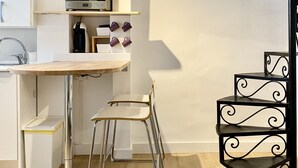 Une cuisine compacte équipée d’un four à micro-ondes, d’une machine à café et d’une petite étagère avec un panier. On y trouve deux chaises blanches et une étagère métallique noire.