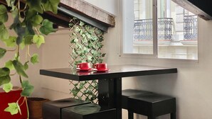 Un coin de pièce avec une table noire, deux tasses rouges posées dessus, un luminaire suspendu en osier et une vue sur un balcon avec des balustrades en fer.