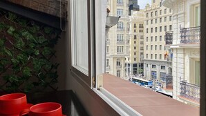Une vue par la fenêtre sur un paysage urbain, avec des immeubles, un balcon et des tasses rouges sur une table.
