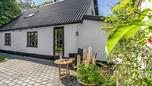 A white building with a black roof, a door, and windows. There is a round table with a chair and a potted plant.