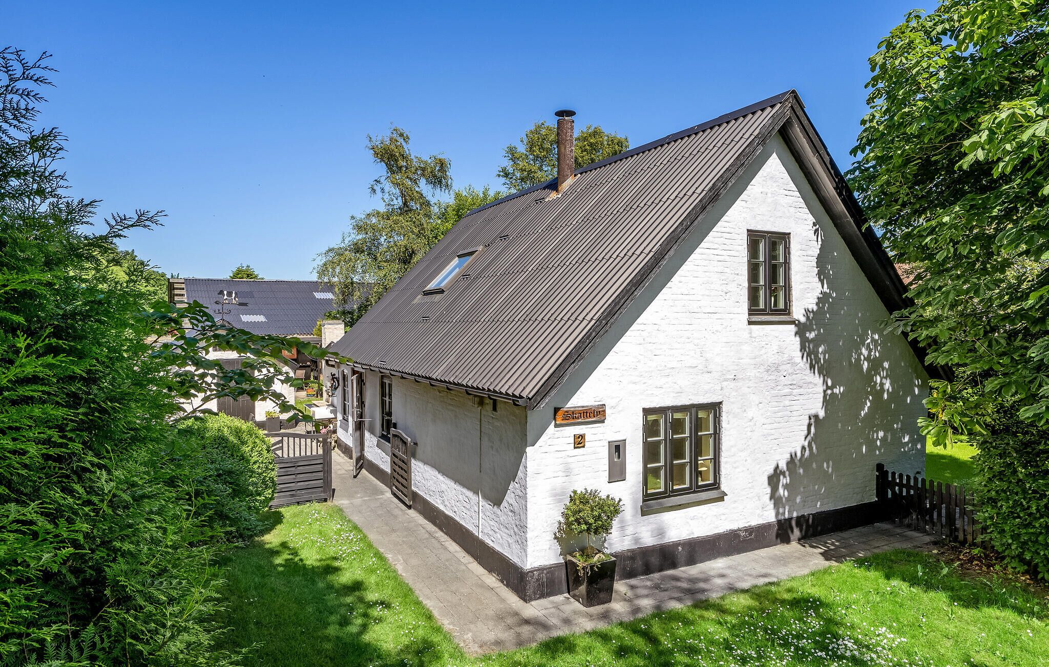 A white house with a sloped roof and a chimney, surrounded by greenery.
