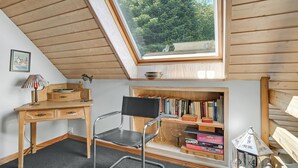 A room with a sloped ceiling, a skylight, a wooden desk, a black chair, a bookshelf, and a small table with a lamp.