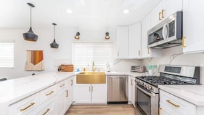 Una cocina moderna con armarios blancos, tiradores dorados, lavavajillas de acero inoxidable, estufa a gas e iluminación colgante.