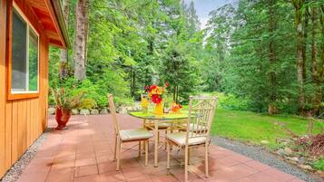 A wooden building with a glass door, a patio with a round table and chairs, and a garden with trees and a lawn.