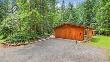 A wooden cabin with a gravel driveway, surrounded by dense forest.