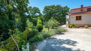 A gravel driveway leading to a house with a tiled roof, surrounded by lush greenery and a swan statue.