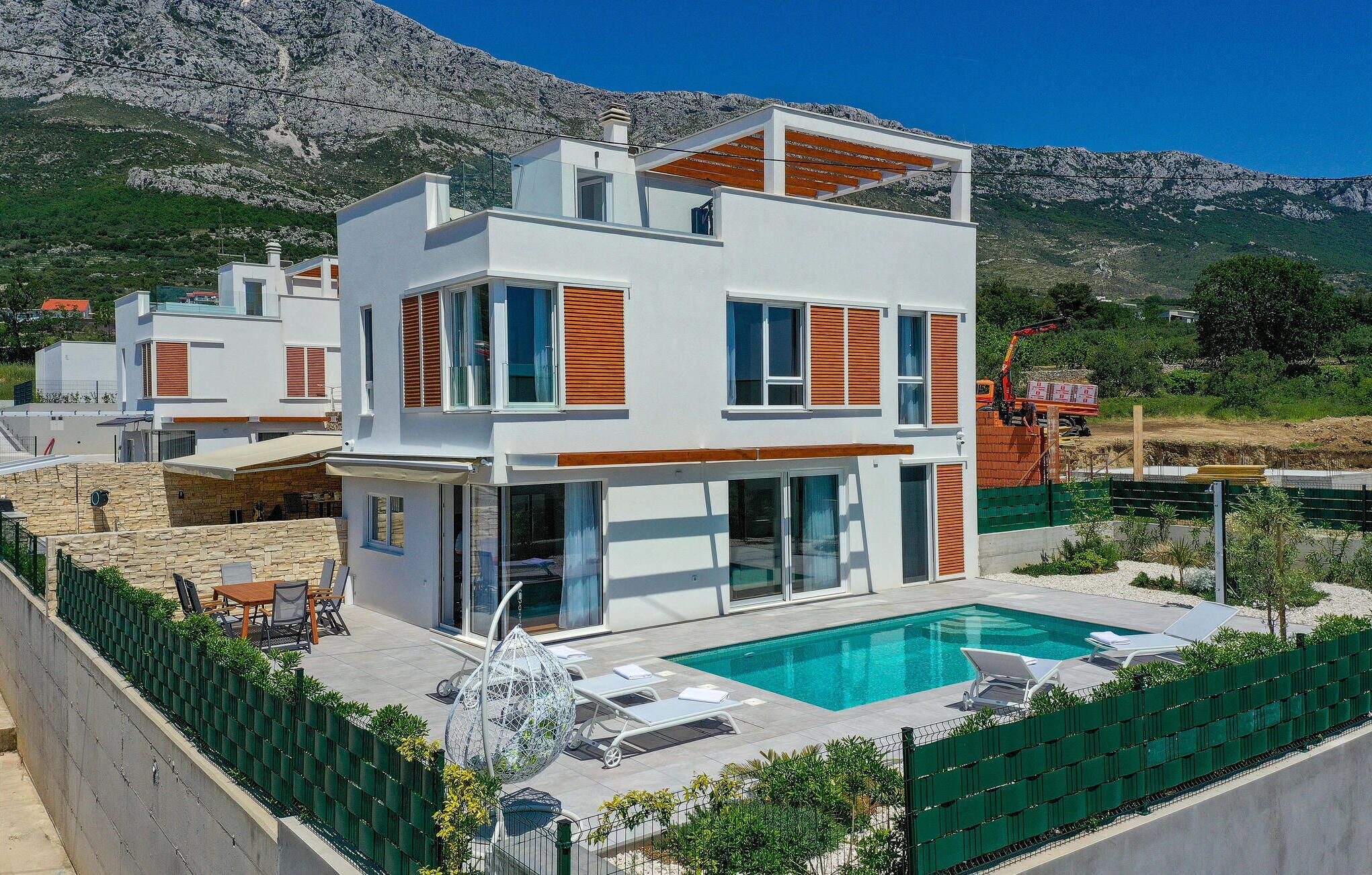 A modern two-story house with a swimming pool, surrounded by a green fence and a mountainous backdrop.