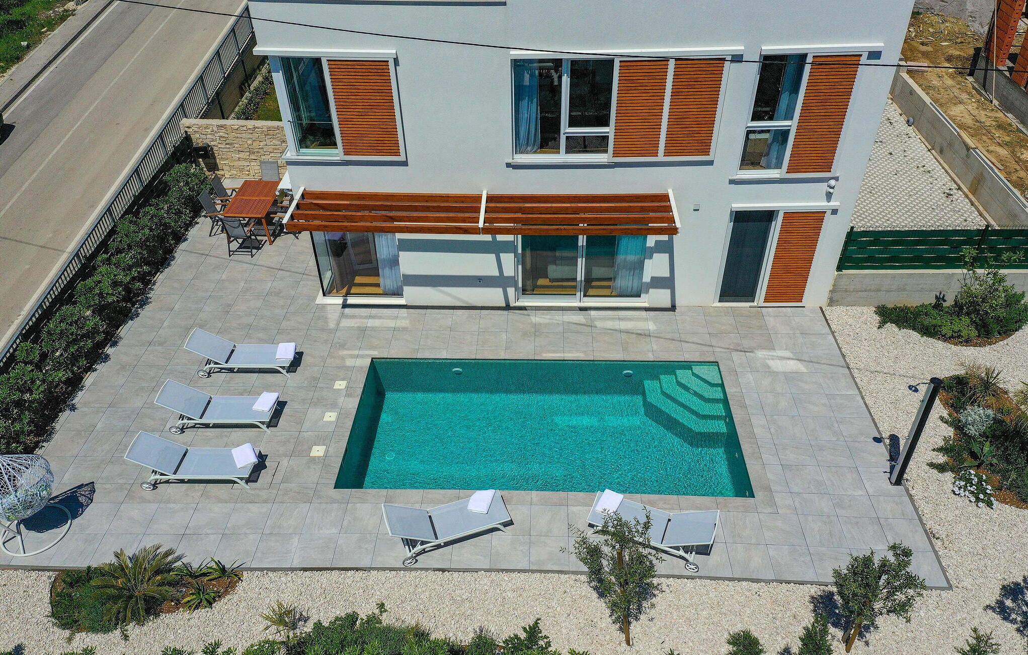 Uma casa moderna de dois andares com piscina retangular, várias espreguiçadeiras e uma pequena área de varanda.