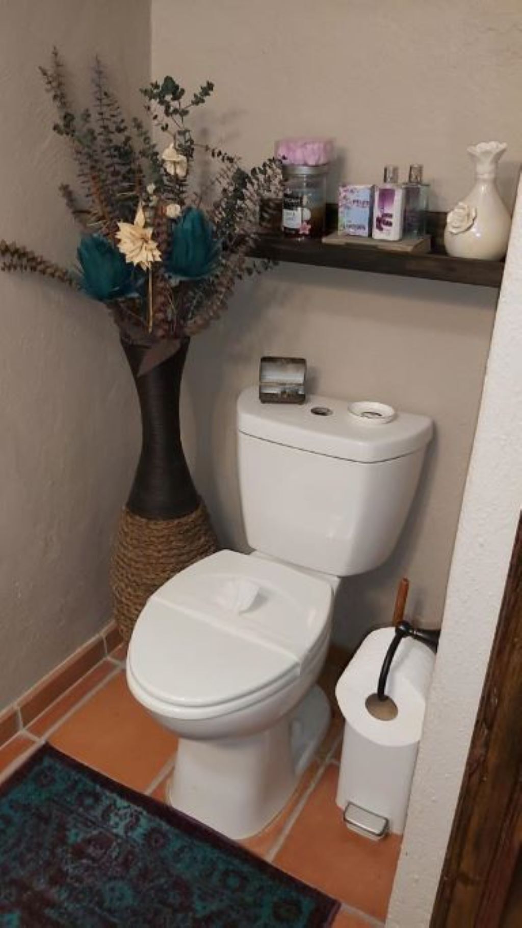 Une salle de bain avec des toilettes blanches, une petite étagère contenant des articles de toilette et un vase décoratif avec des fleurs.