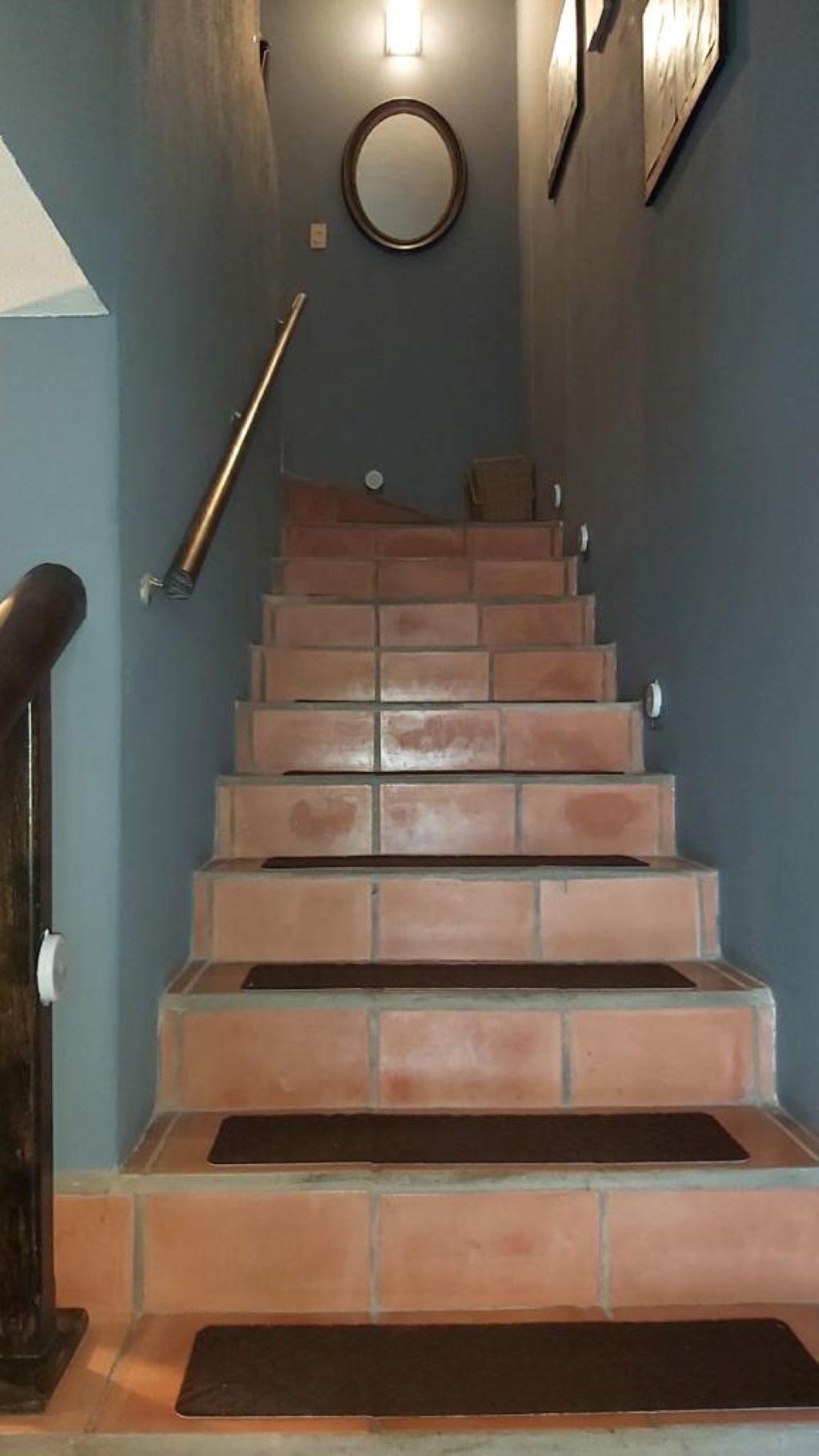 A staircase with terracotta steps and a handrail, leading to a landing with a round mirror and wall-mounted light fixtures.