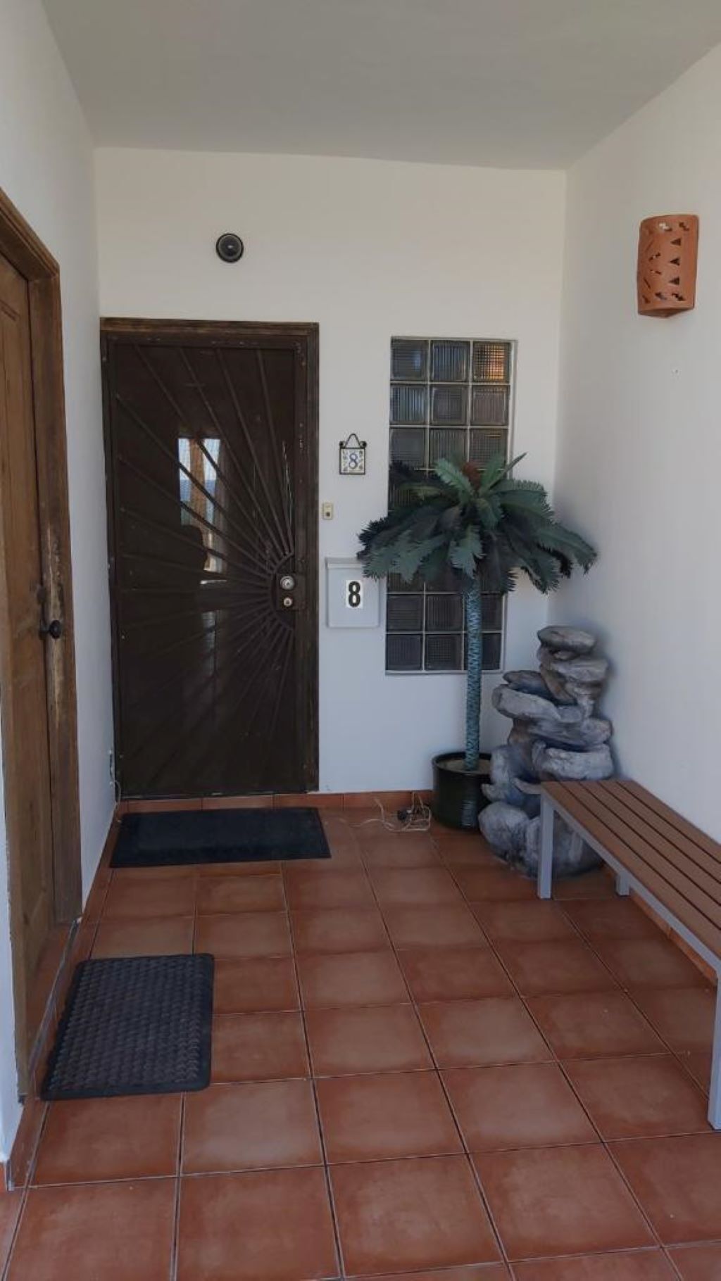 Un hall d’entrée carrelé avec une porte en bois, une plante en pot, un banc et une petite fenêtre sur laquelle est apposé le numéro « 8 ».