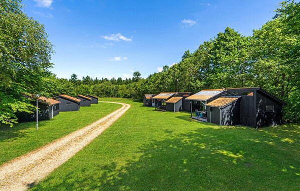 A row of small cabins with sloped roofs, a gravel path leading through the grassy area, and dense trees in the background.