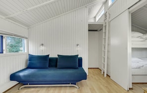 Quarto com sofá azul, duas luzes fixadas na parede, janela com persianas azuis e uma cama beliche com roupa de cama branca.