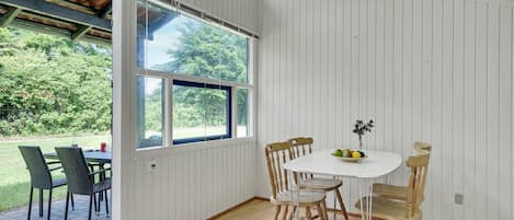 Et værelse med en hvid panelvæg, et træbord med stole og et stort vindue med udsigt til grøn natur.